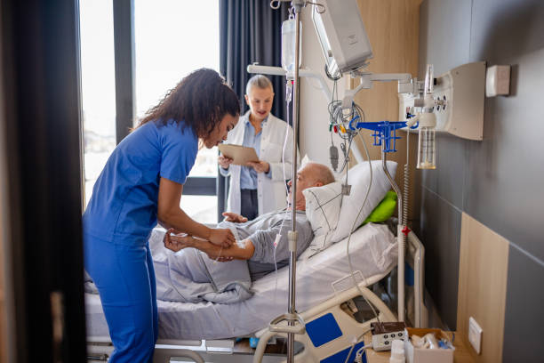 A senior man lies in a hospital bed as a nurse provides care while a doctor reviews medical notes. The setting showcases a healthcare environment focused on patient support.